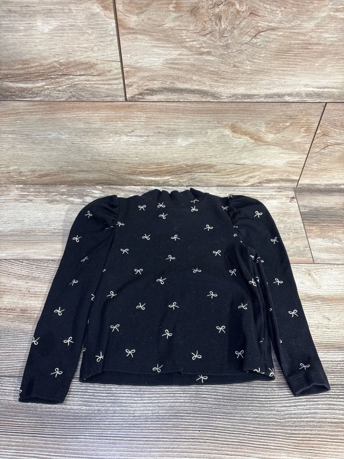 Front view of Baby Gap black ribbed mockneck shirt in size 3T showcasing puff sleeves and bow print on wood floor