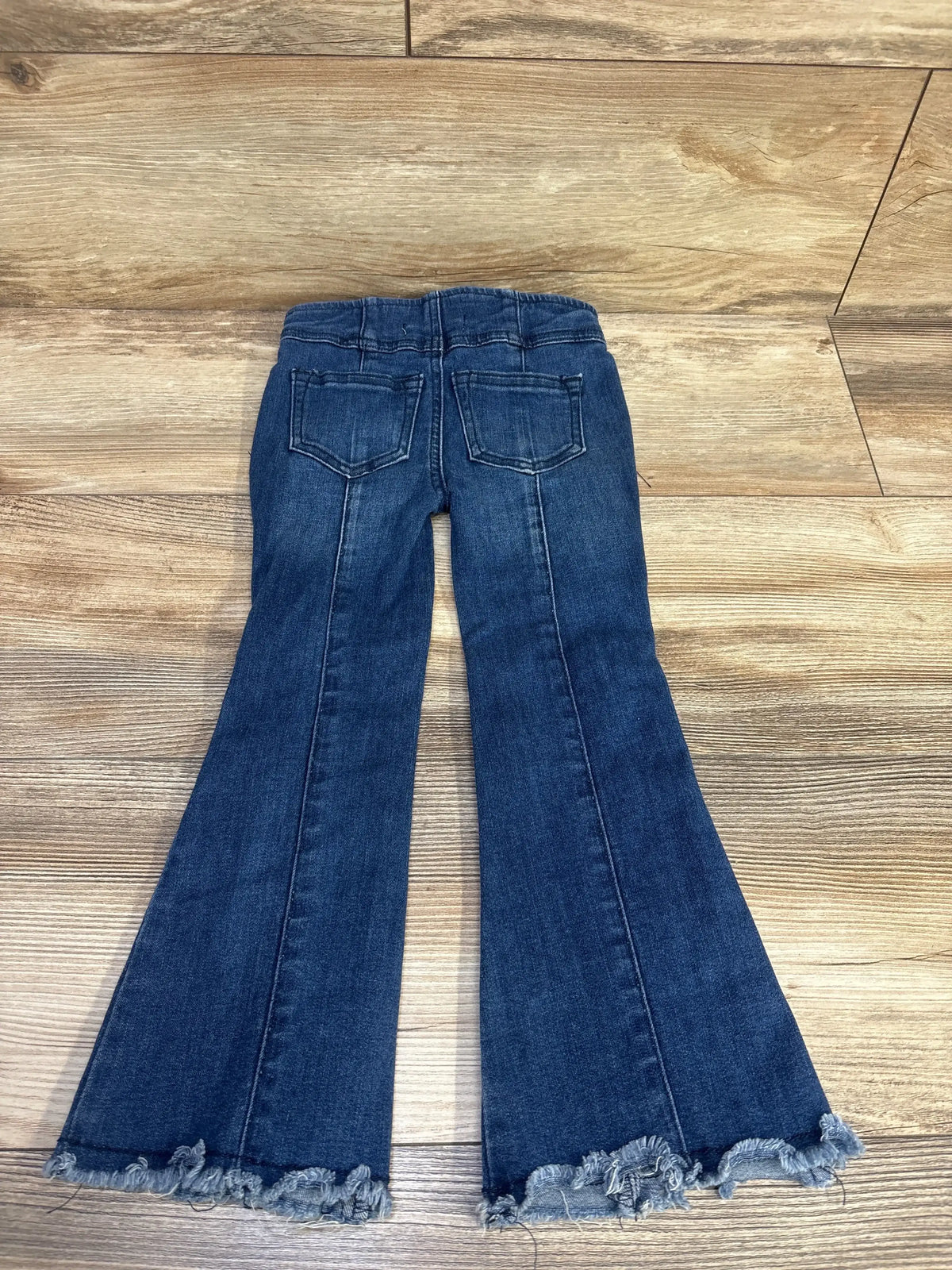 Back view of Chelsea & Violet blue flare jeans with frayed hems and back pockets, laid flat on wood background