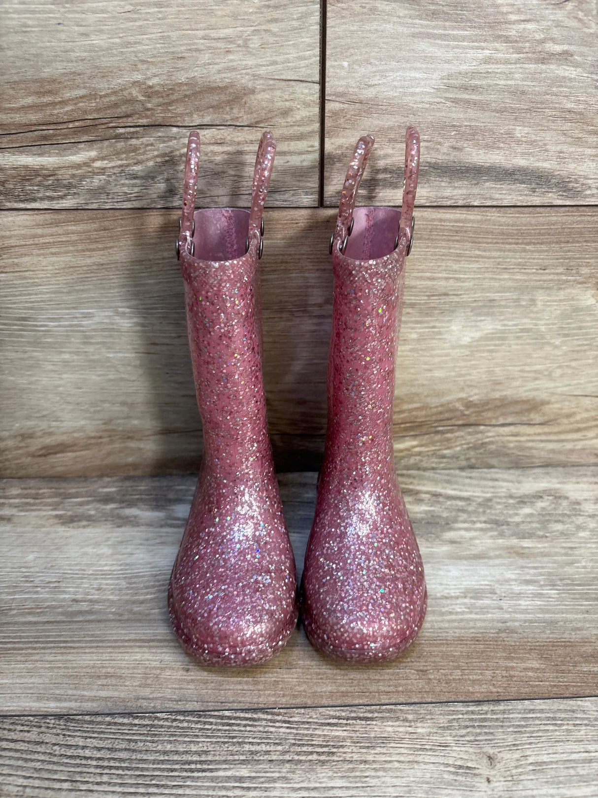 Front view of Western Chief Abby light-up pink glitter kids rain boots showcasing glitter finish and pull handles on a wood floor