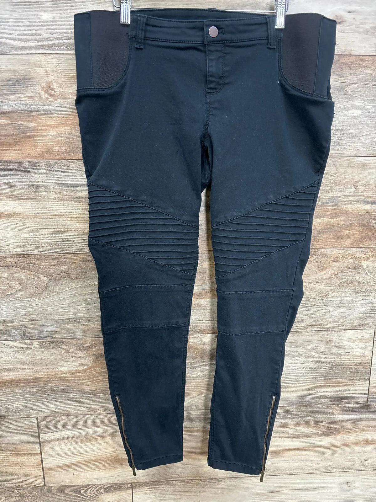 Black moto-style maternity pants with ribbed knee panels and ankle zippers, flat lay on wood