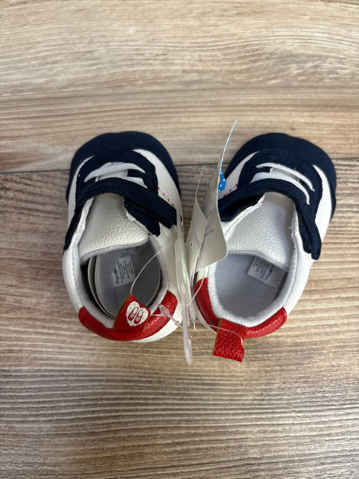 Overhead view of Stepping Stones baby baseball shoes, size 9–12 months, white with navy straps and red heel tabs on wood background