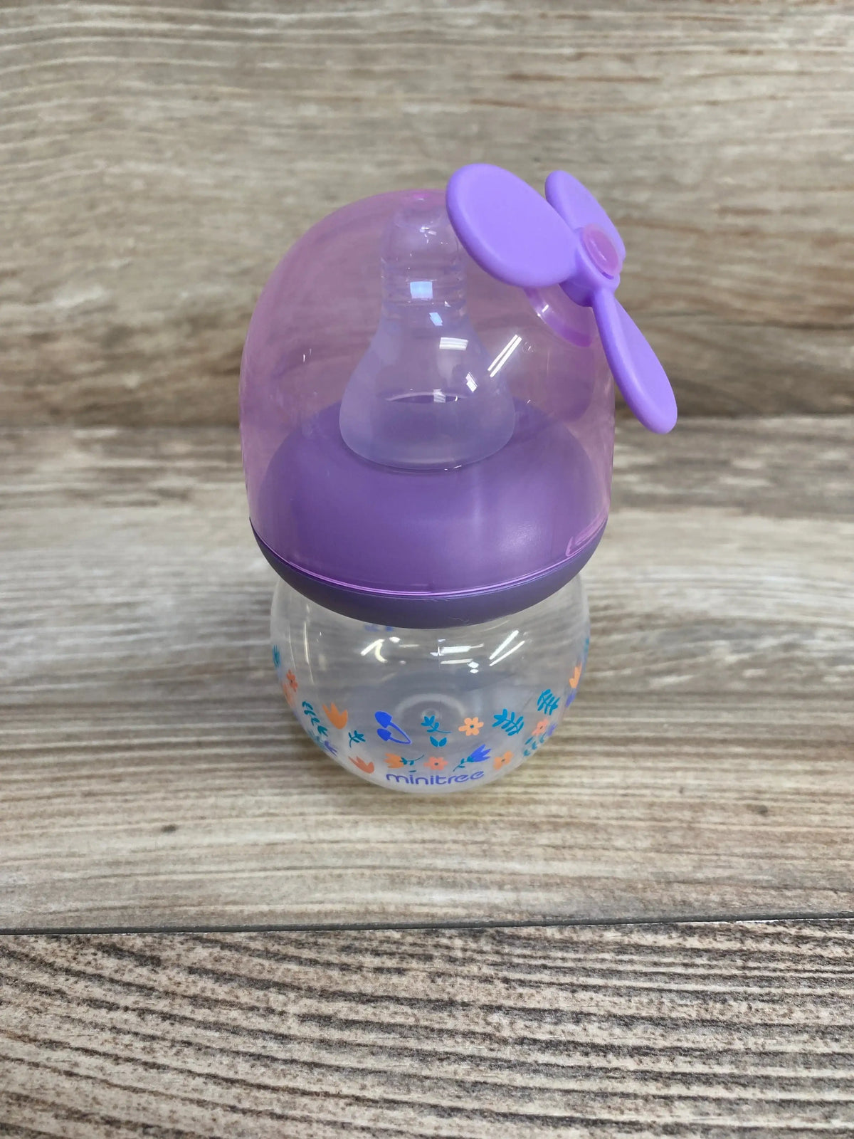 Front view of MINITree 2oz purple baby bottle with nipple, dome lid and propeller cap, floral print on wood background