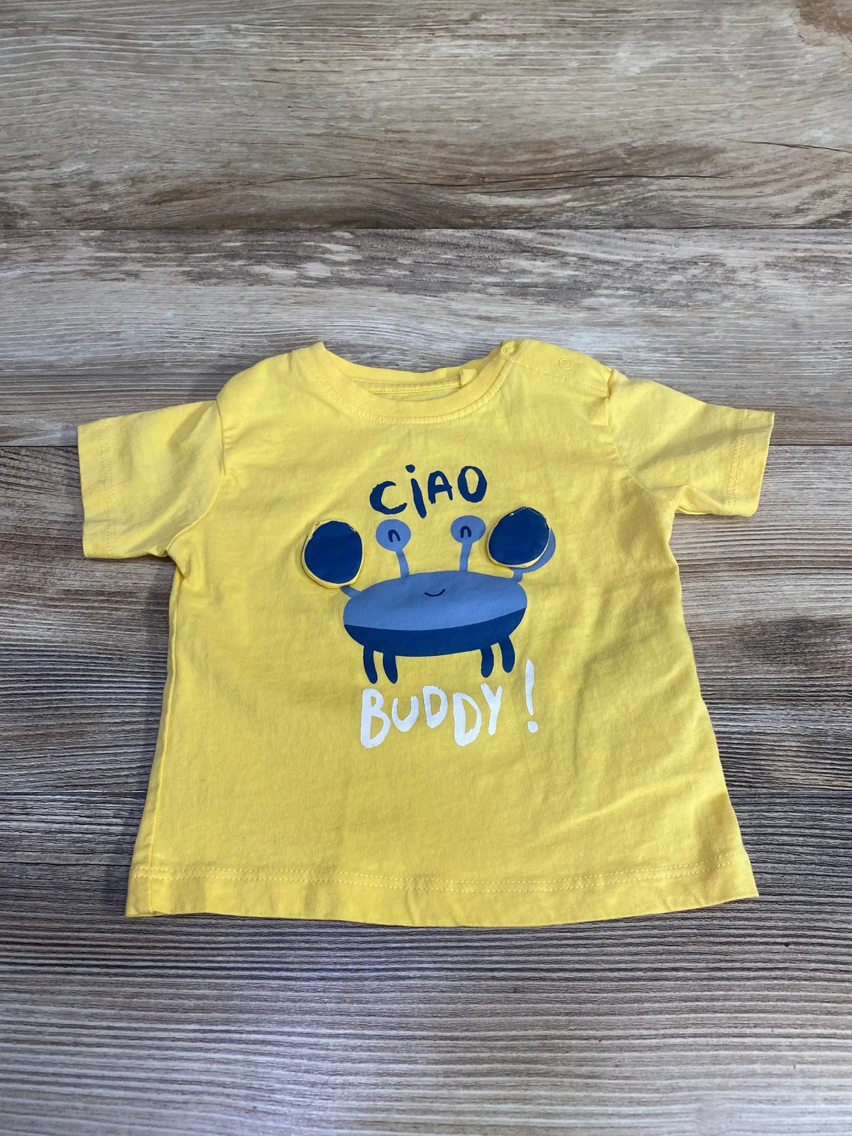 Flat lay of MO Kids Ciao Buddy shirt sz 6-9m showcasing yellow tee with blue crab graphic and Ciao Buddy lettering on wood plank floor