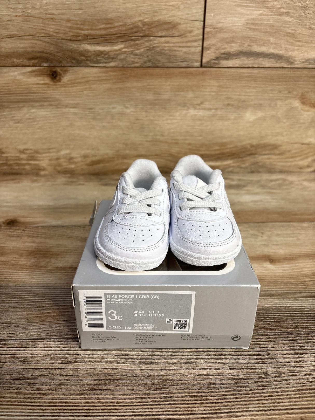 White Nike Force 1 Crib baby shoes on gray box with wood background, size 3C