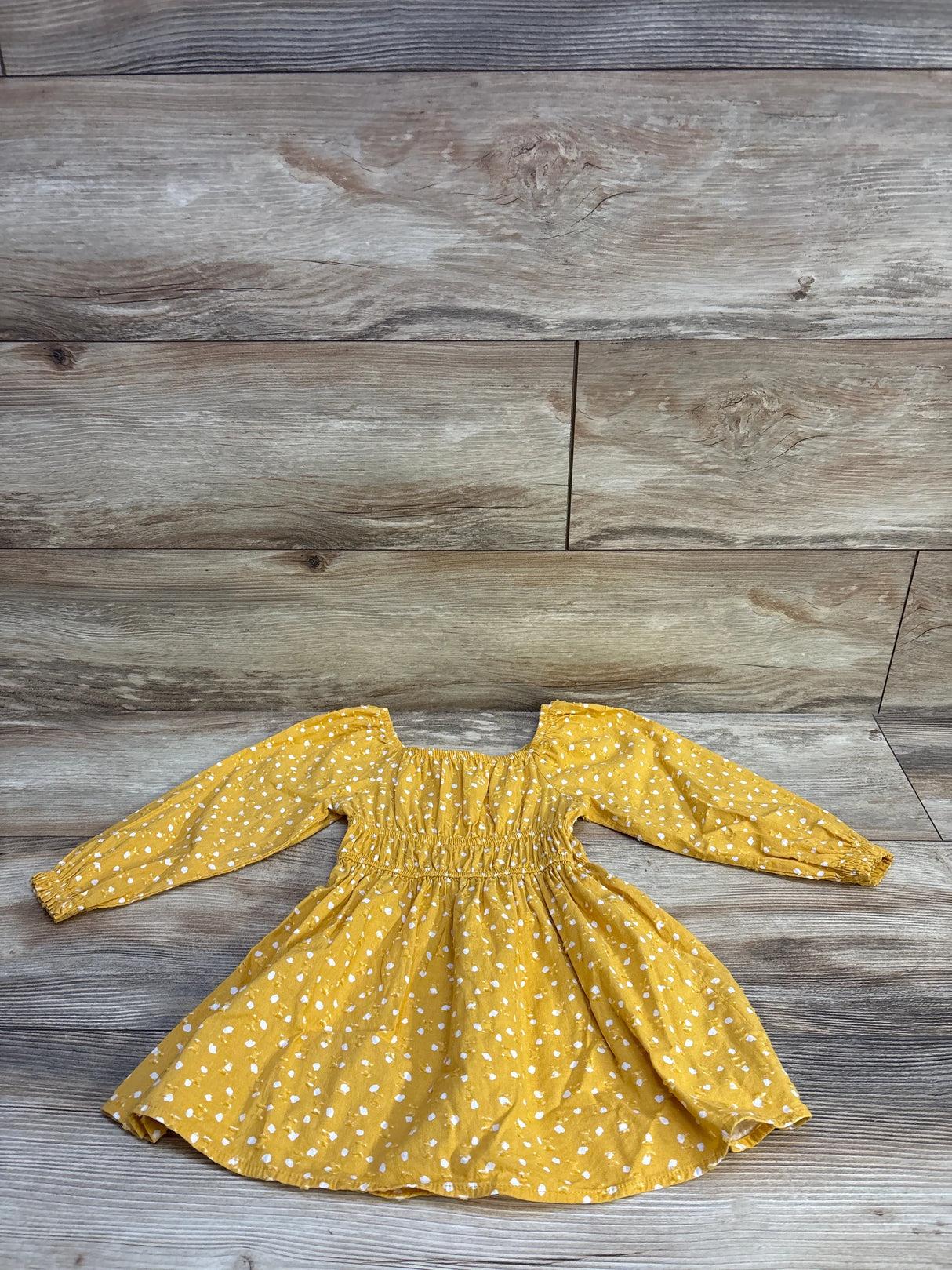 Flat lay of yellow long-sleeve Swiss dot toddler dress showcasing smocked bodice and elastic cuffs on wood plank background