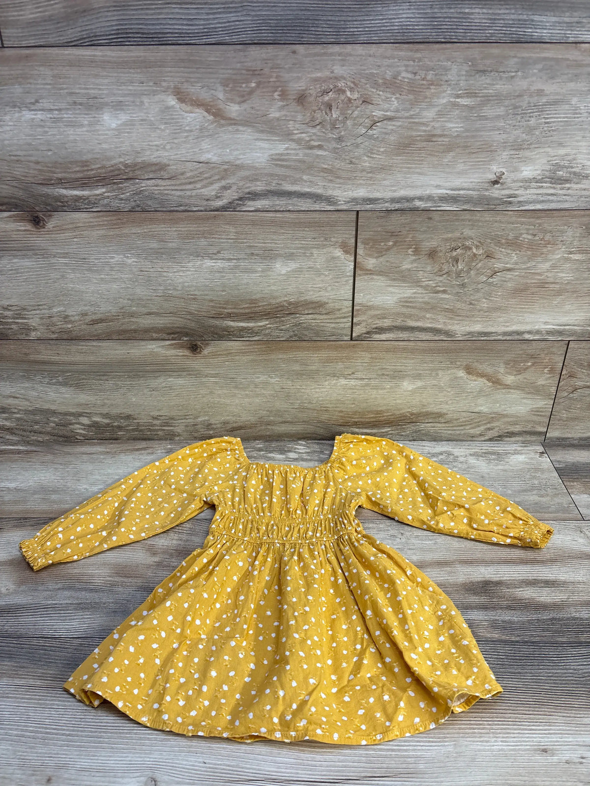Flat lay of yellow long-sleeve Swiss dot toddler dress showcasing smocked bodice and elastic cuffs on wood plank background