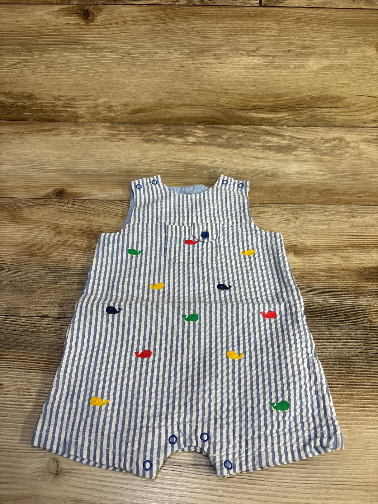 Front flat-lay of baby shortie romper showing blue-and-white seersucker stripes and small multicolored embroidered whales on wooden floor