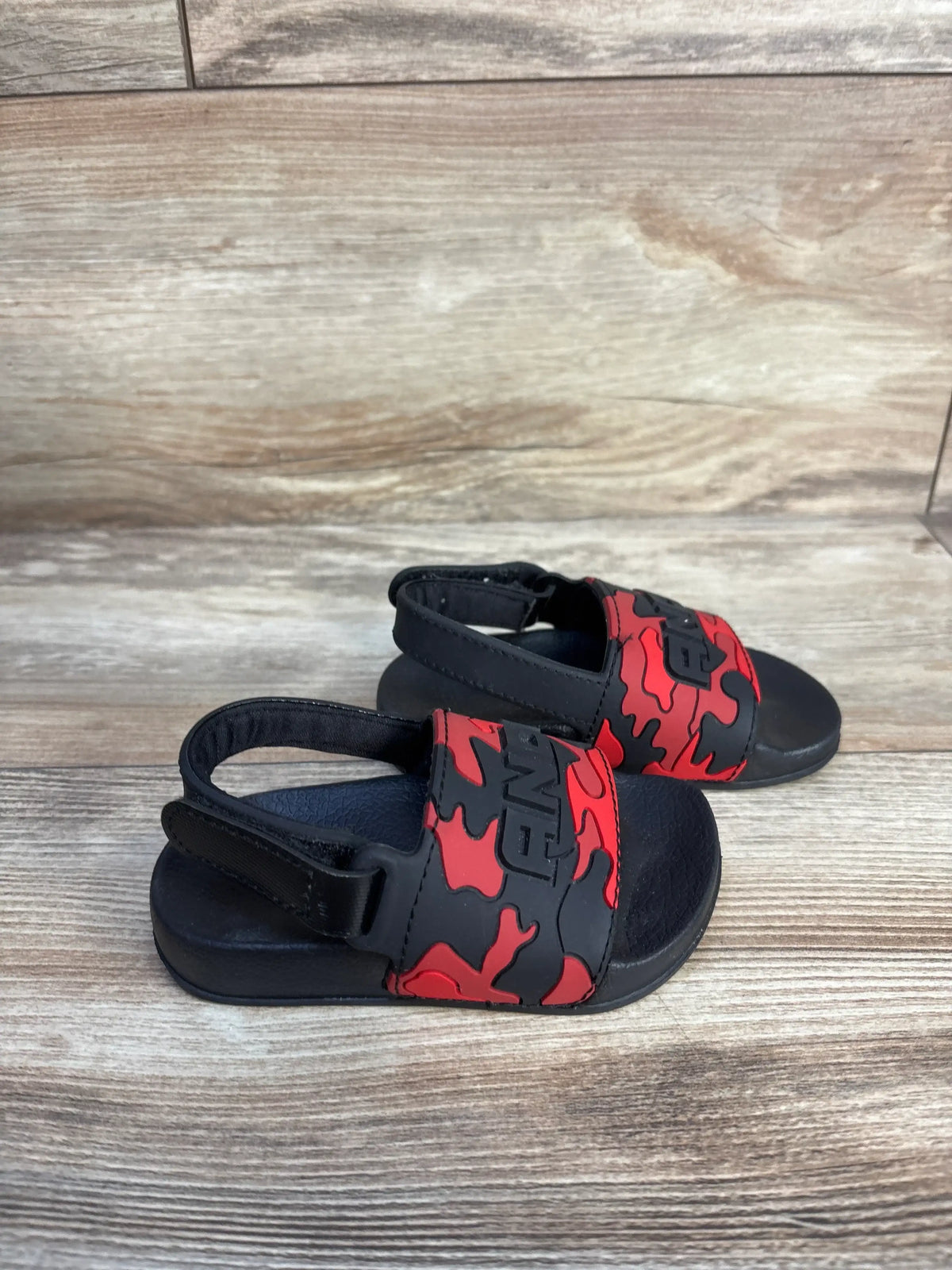 Angled view of And1 camo slide sandals showcasing red and black camouflage straps and black soles on a wood-grain background