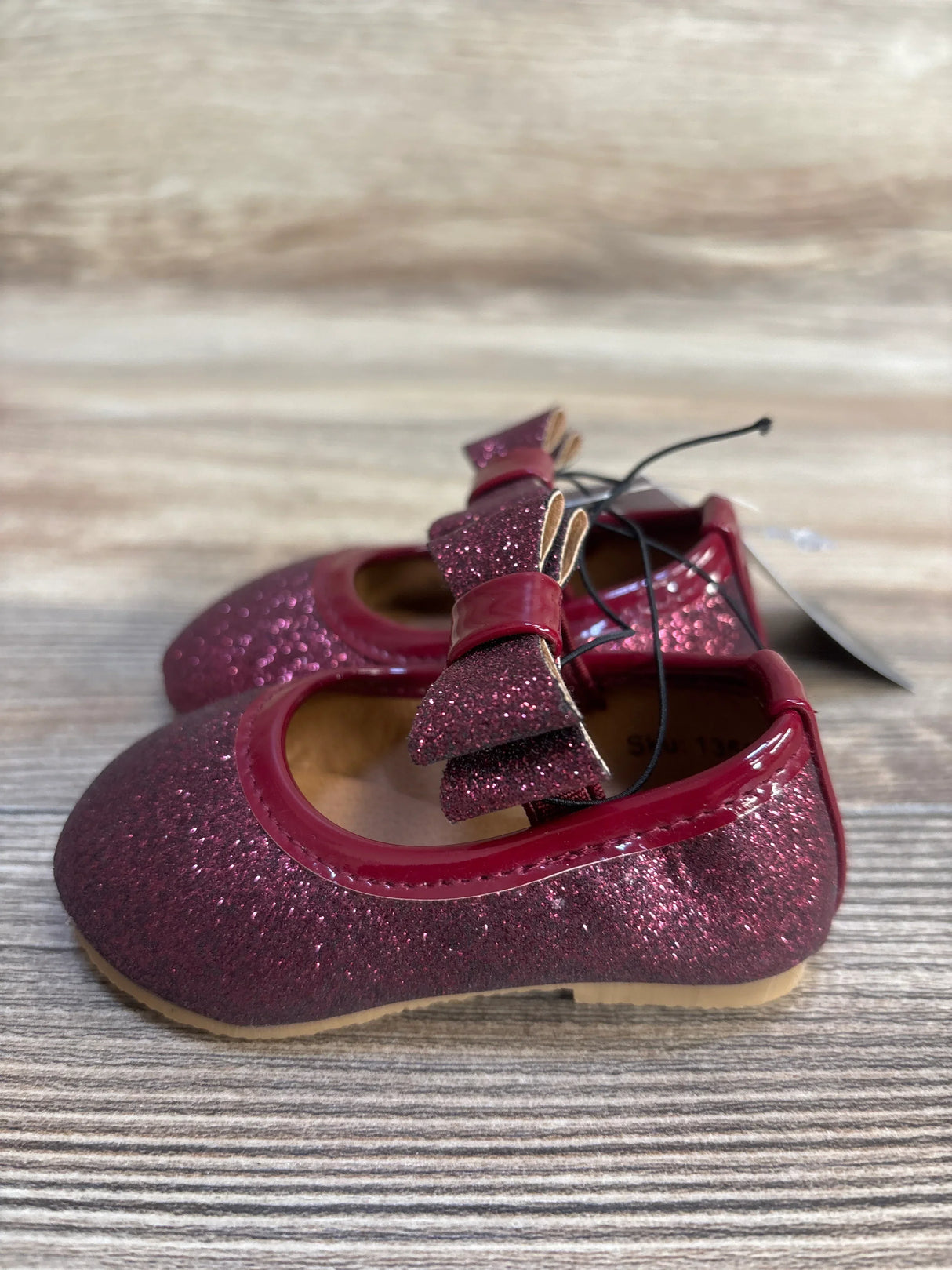 Side view of maroon Mary Jane baby flats with glitter finish and bow detail on light wood surface