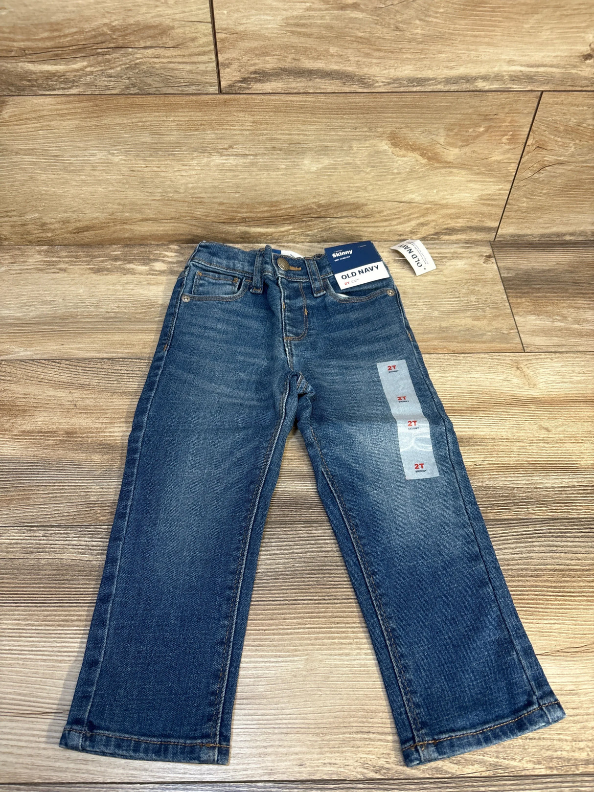 Front view of Old Navy toddler skinny jeans size 2T showing faded wash, attached tags, and size stickers on wood floor background