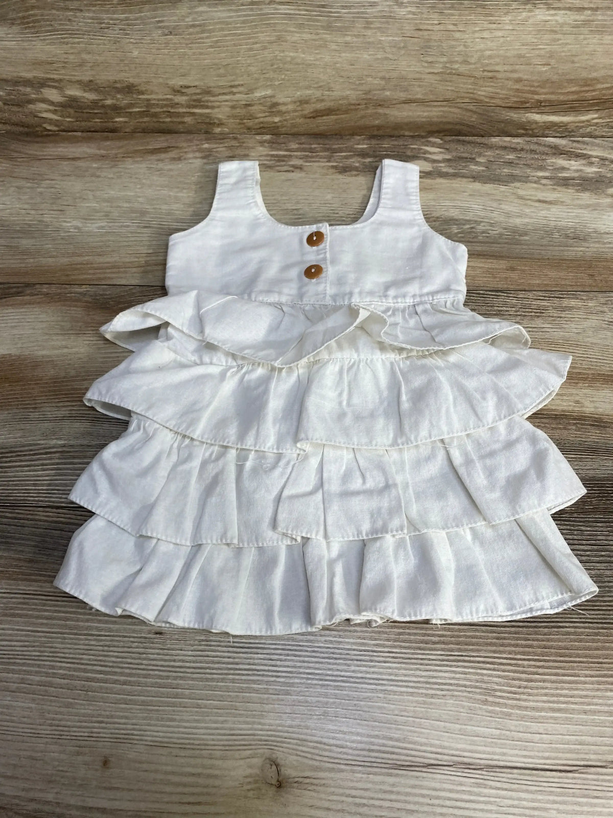Top-down view of white tiered baby dress with three ruffled layers and two wooden buttons on a light wood background