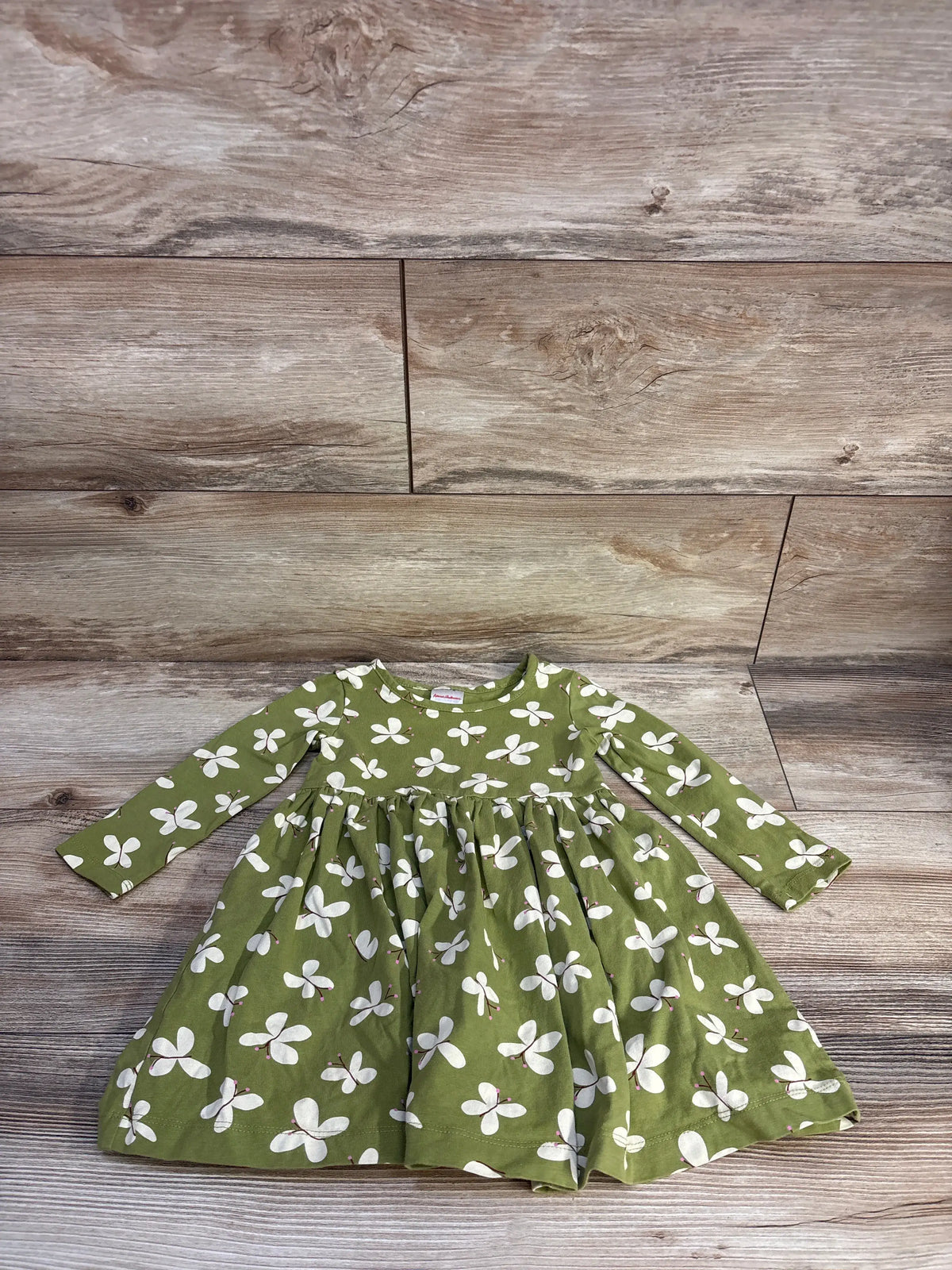 Flat lay of Hanna Andersson long-sleeve skater dress showcasing moss green fabric with white butterfly print on wooden floor background, size 2T