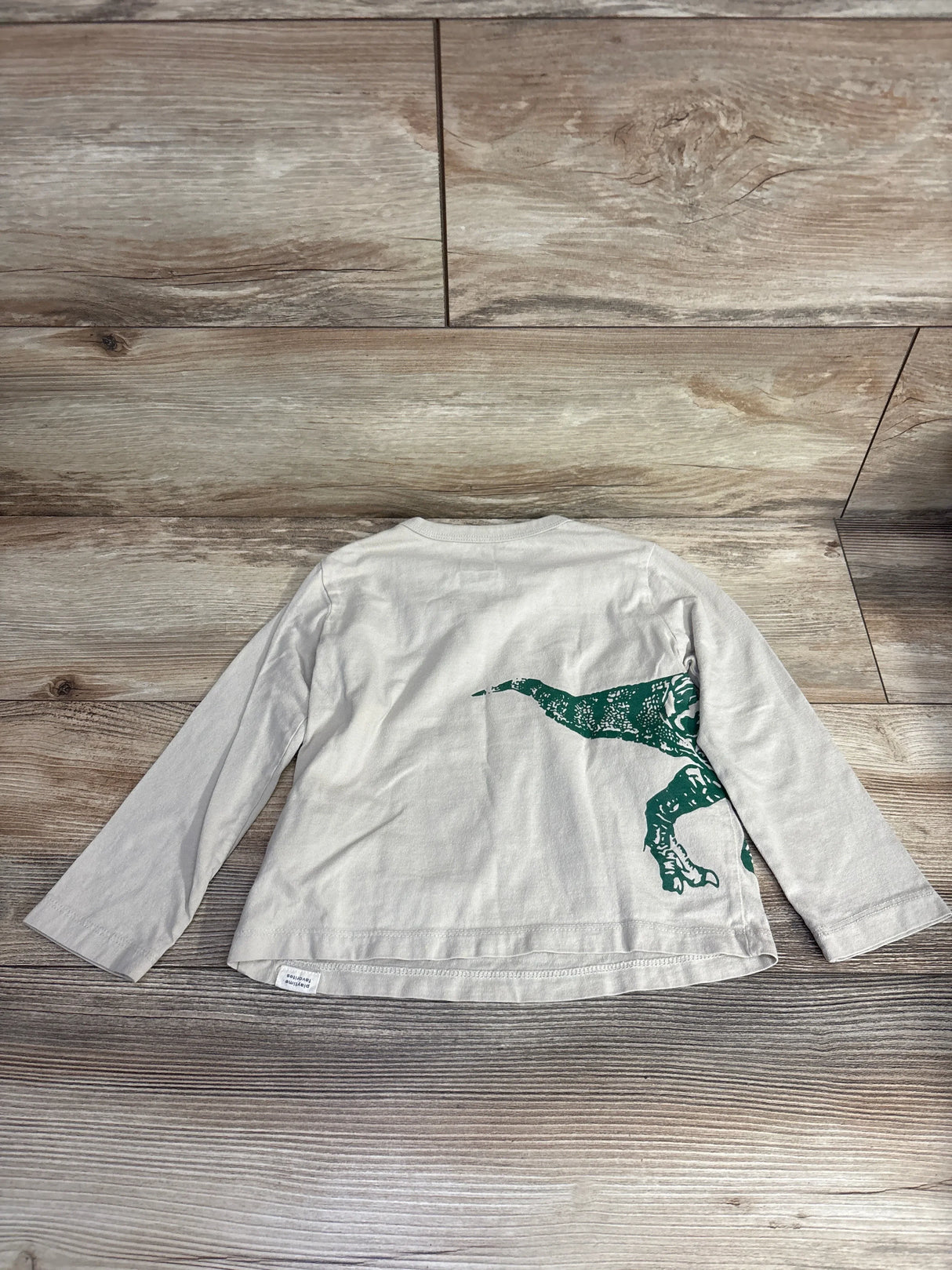 Beige long sleeve kids shirt with green dinosaur graphic on wooden floor