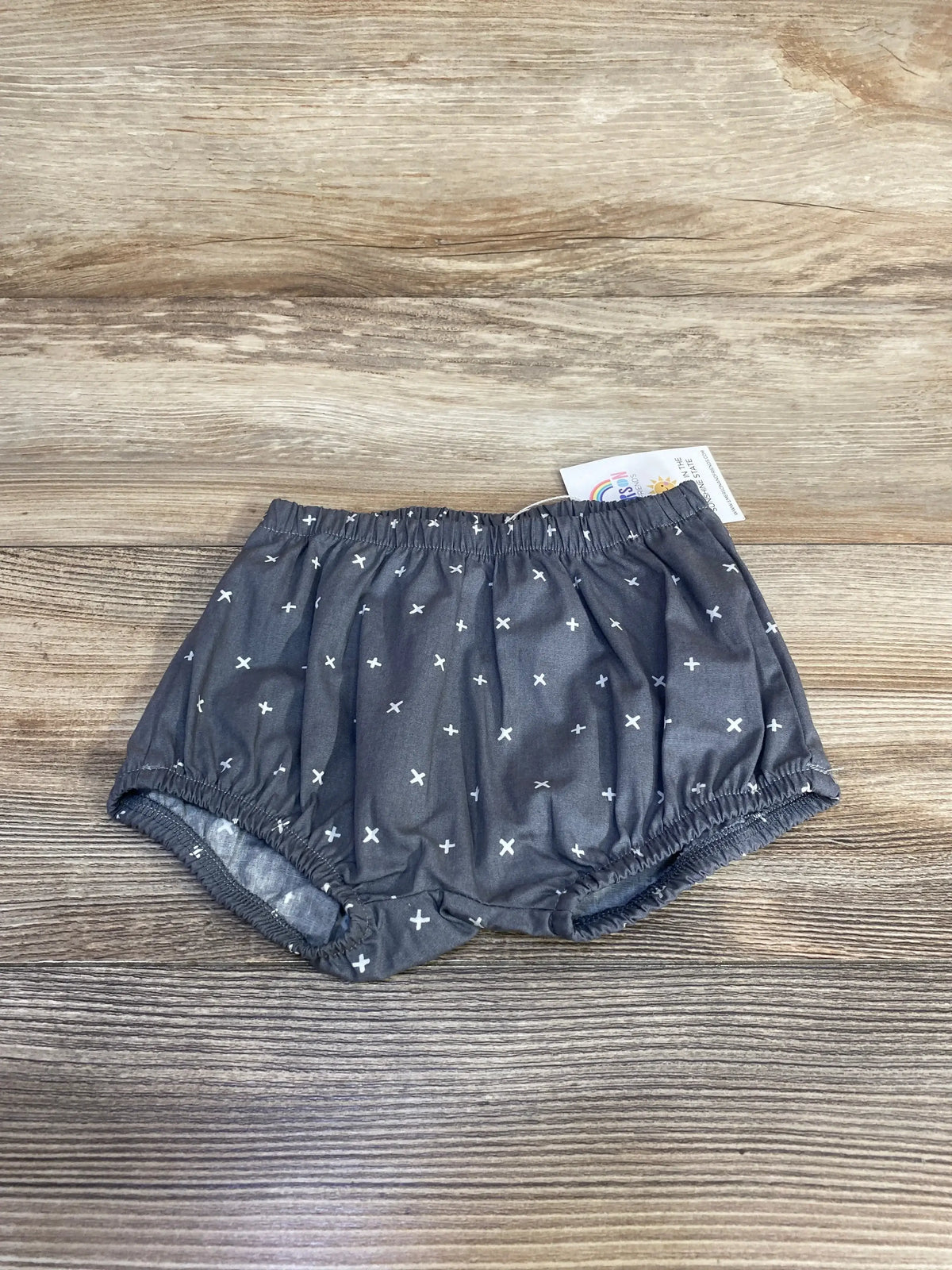 Top-down view of grey baby bloomers size 12 to 18 months with elastic waist and white cross print on wood backdrop