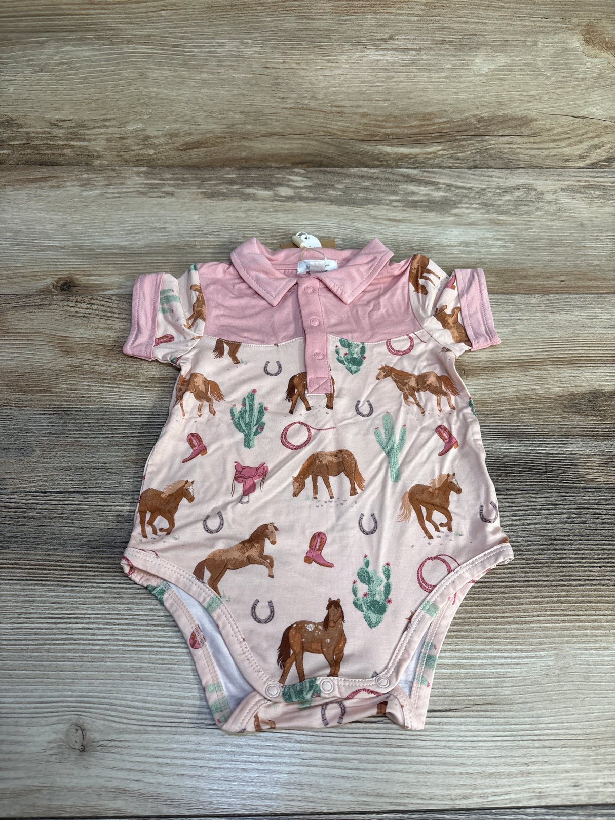 Pink baby onesie with horses, cacti, cowboy boots, and horseshoes on wood background