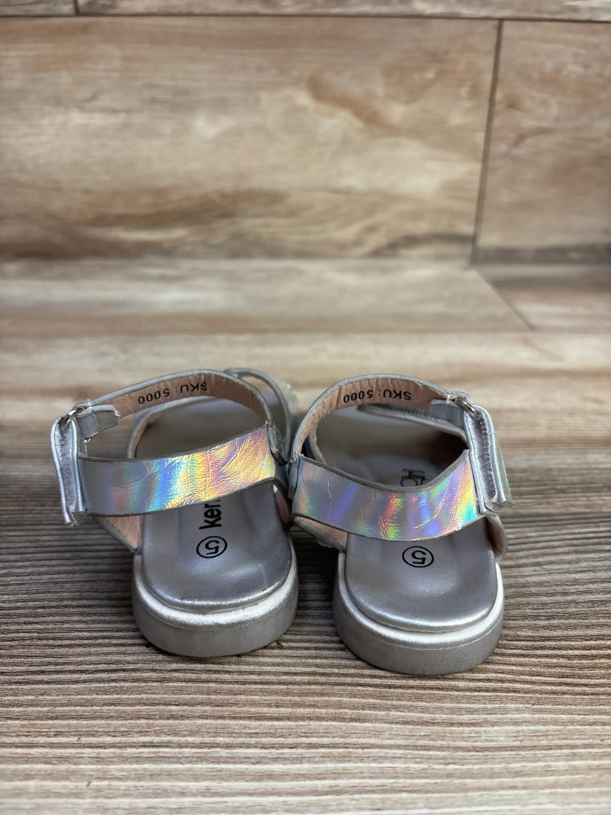 Front view of Kensie Girl glitter sandals in silver, showcasing glitter finish and hook and loop ankle straps on wood floor