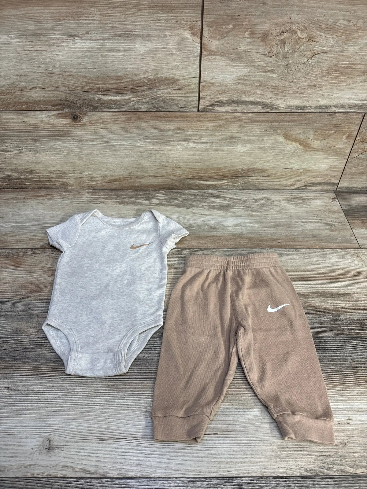 Top-down view of Nike baby two-piece 6m showing gray bodysuit with small swoosh and tan cuffed pants on wood floor