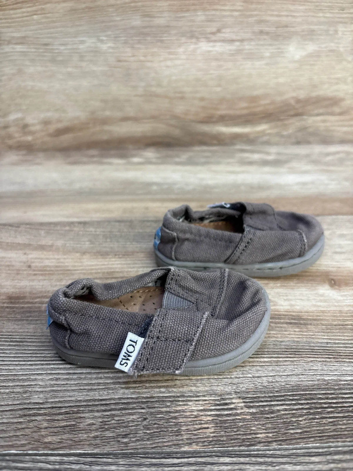 Side view of Toms Tiny Alpargata shoes in grey with canvas upper, elastic gore and TOMS tag on wood-grain background