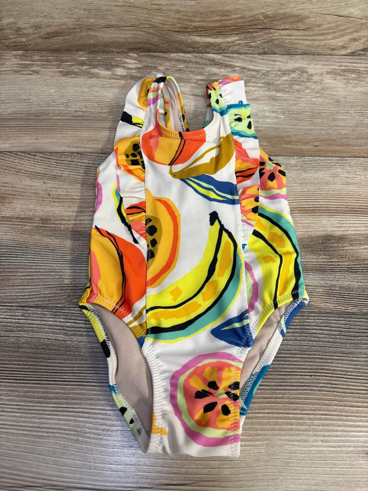 Flat lay of baby one-piece swimsuit with ruffle shoulder straps and colorful fruit print on wood background, 12-18 months