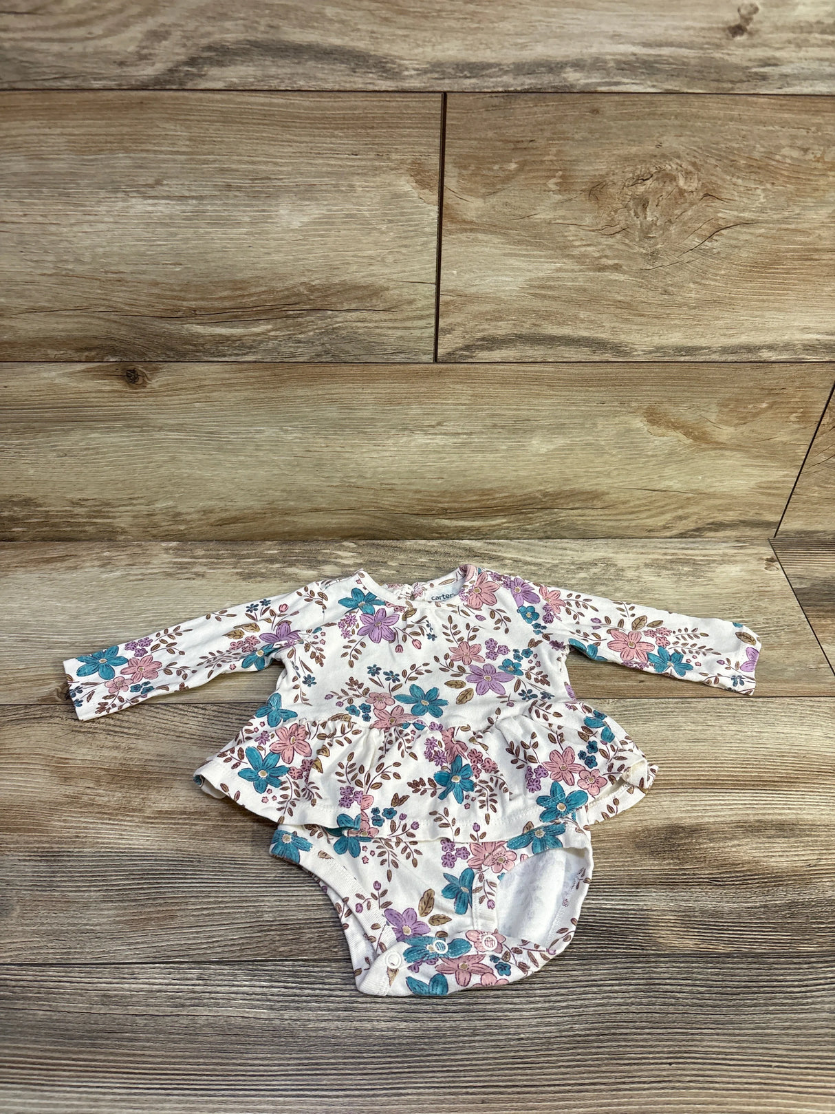 Flat lay of Carter’s floral skirted bodysuit, size 6 months, showcasing ruffled skirt and snap crotch with light wood background
