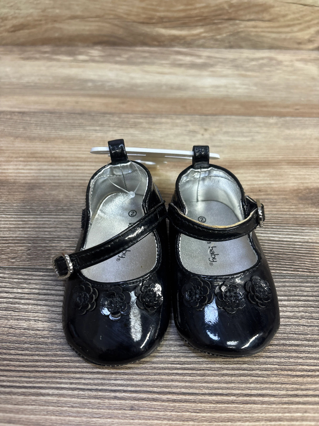 Top-down view of Koala baby black patent Mary Jane flats showcasing three floral appliques and adjustable buckle strap on a wood-grain background