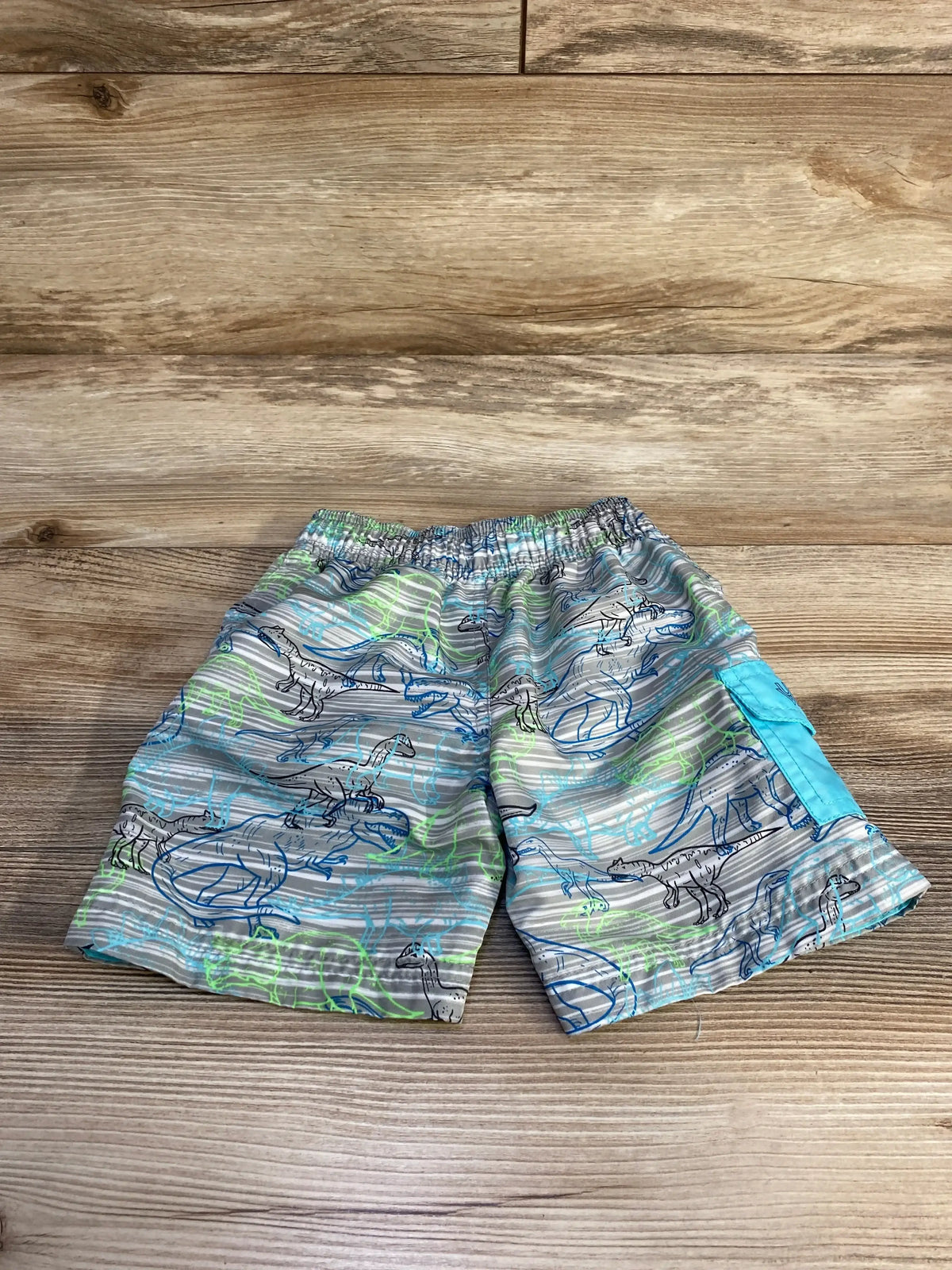 Flat-lay UV Skinz toddler swim trunks size 2T, grey dinosaur print with turquoise side pocket on wooden background