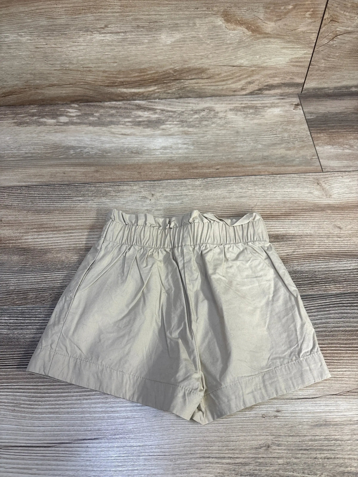 Top-down view of Janie & Jack beige baby shorts (6-12 months) showing elastic ruffle waistband and side pockets on wood floor background