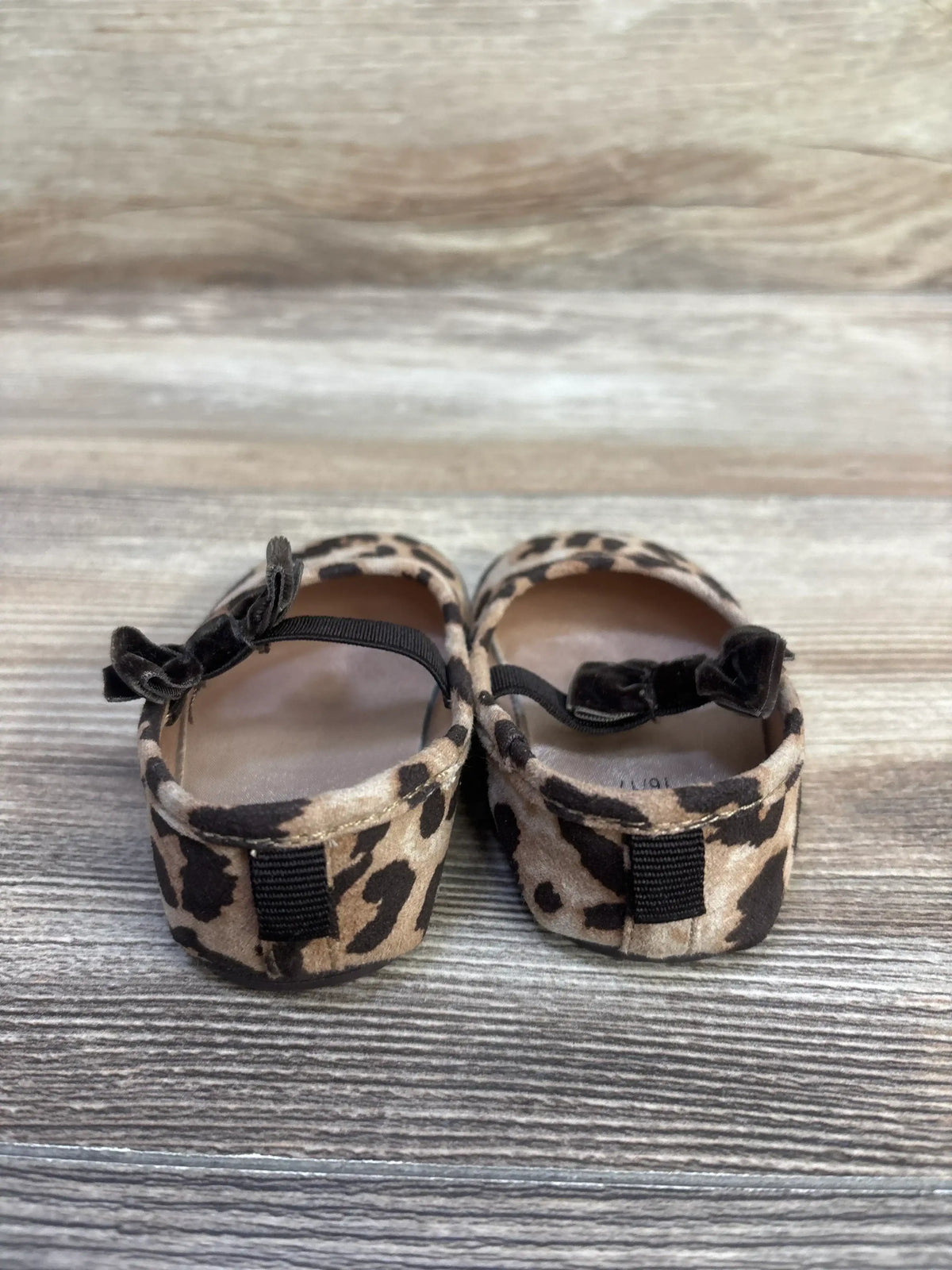 Rear view of H&M leopard-print toddler ballet flats showcasing velvet bow straps and elastic ankle bands on a wood floor background