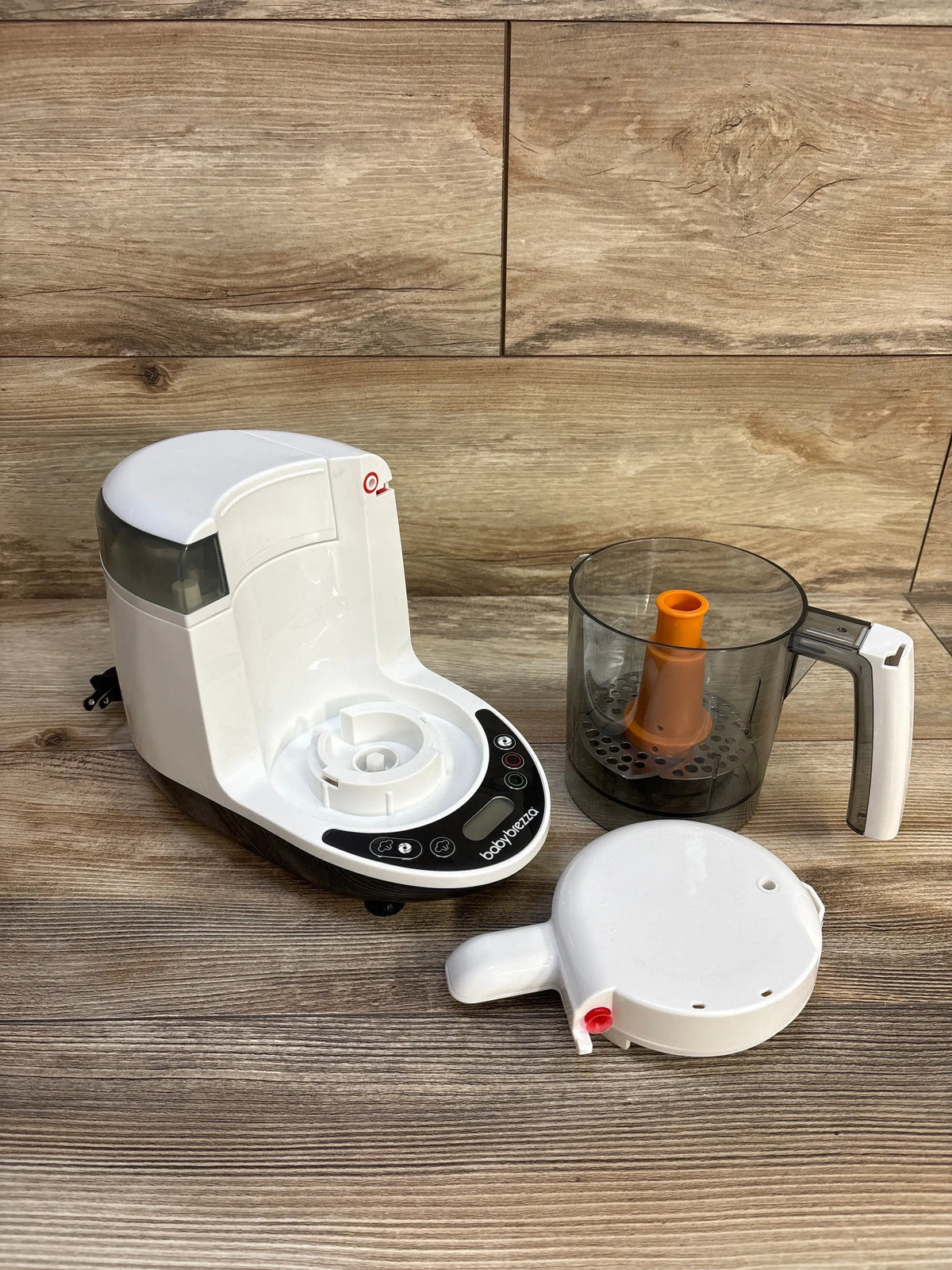 Angled view of Baby Brezza One Step Baby Food Maker Deluxe showcasing white base, clear food bowl, orange blade, and detached lid