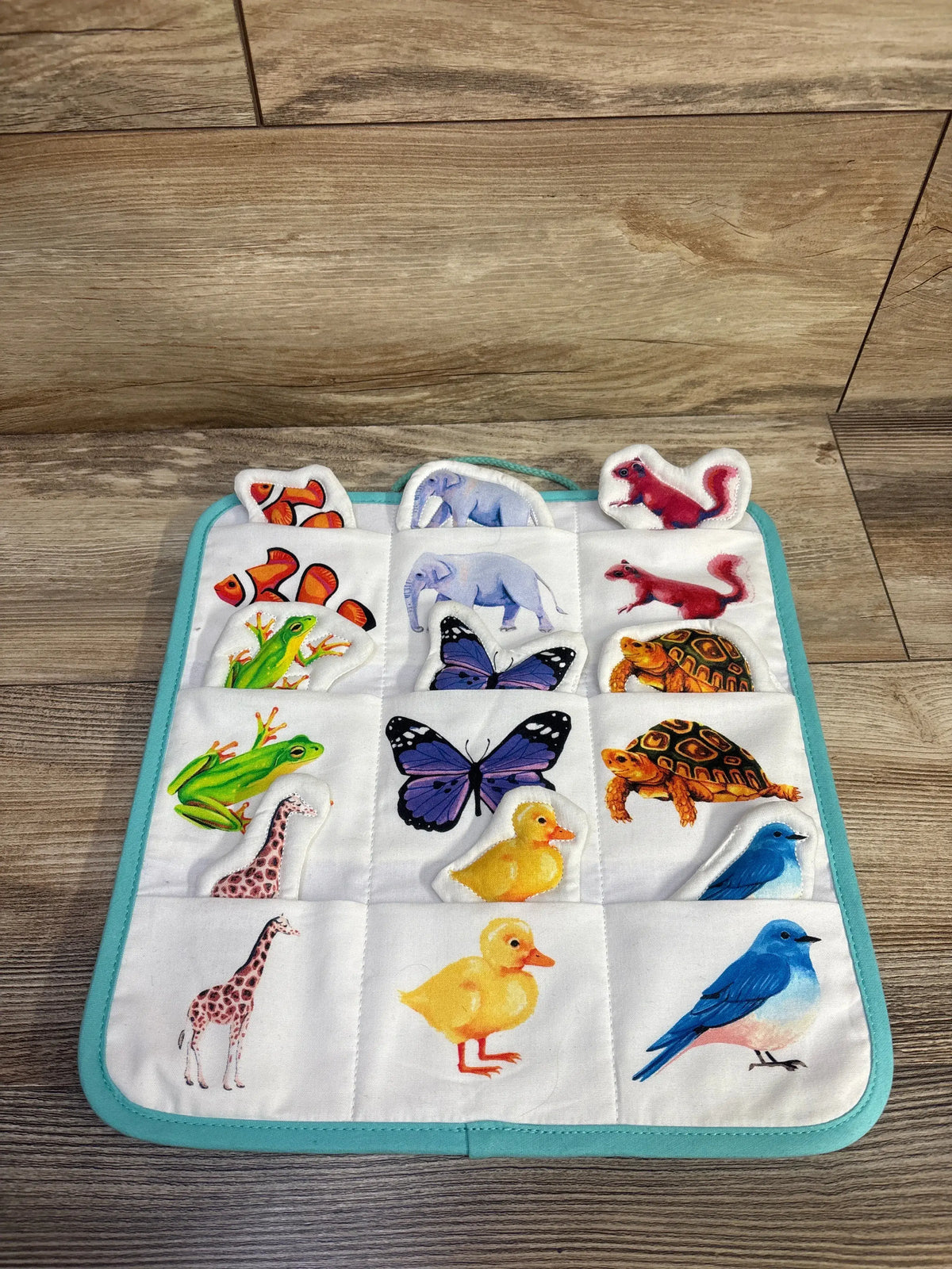 Front view of Lovevery Quilted Critter Pockets showcasing animal-shaped cotton pieces and turquoise trim on a wood floor