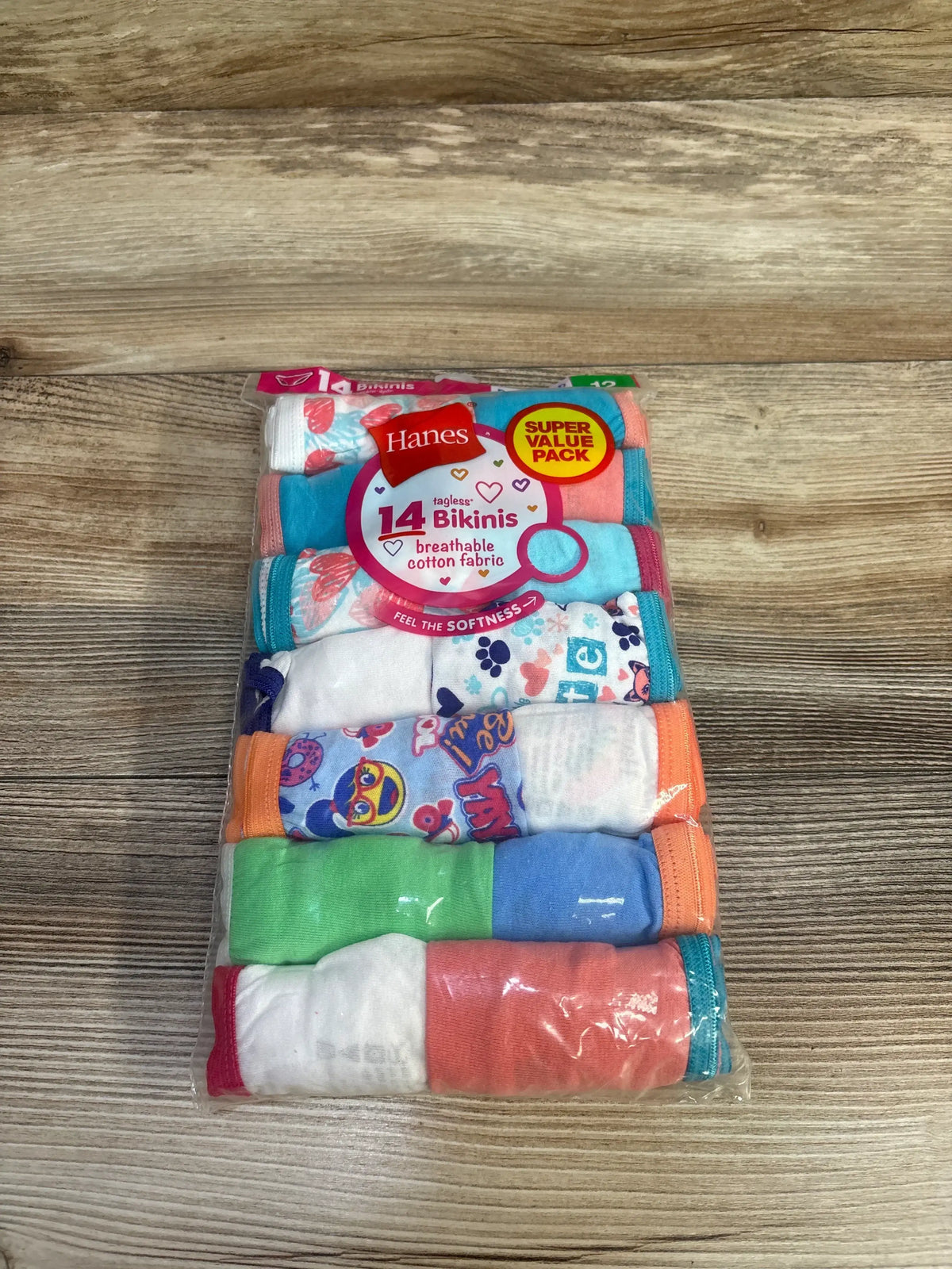 Top-down view of Hanes 14-pack girls’ bikini underwear showing assorted colorful solids and patterned pairs in clear plastic packaging on a wooden surface