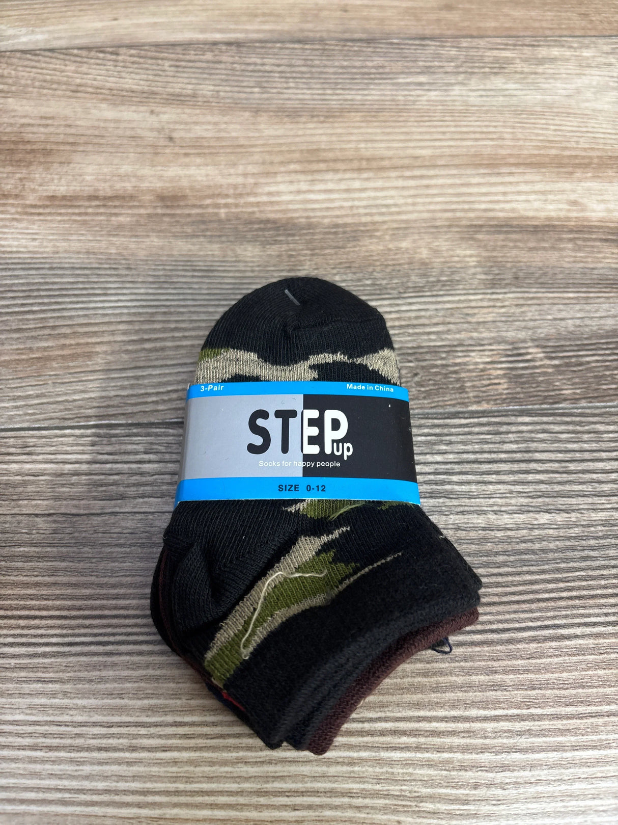 Three pairs of black and camouflage baby socks labeled Step Up, size 0-12, on wooden surface