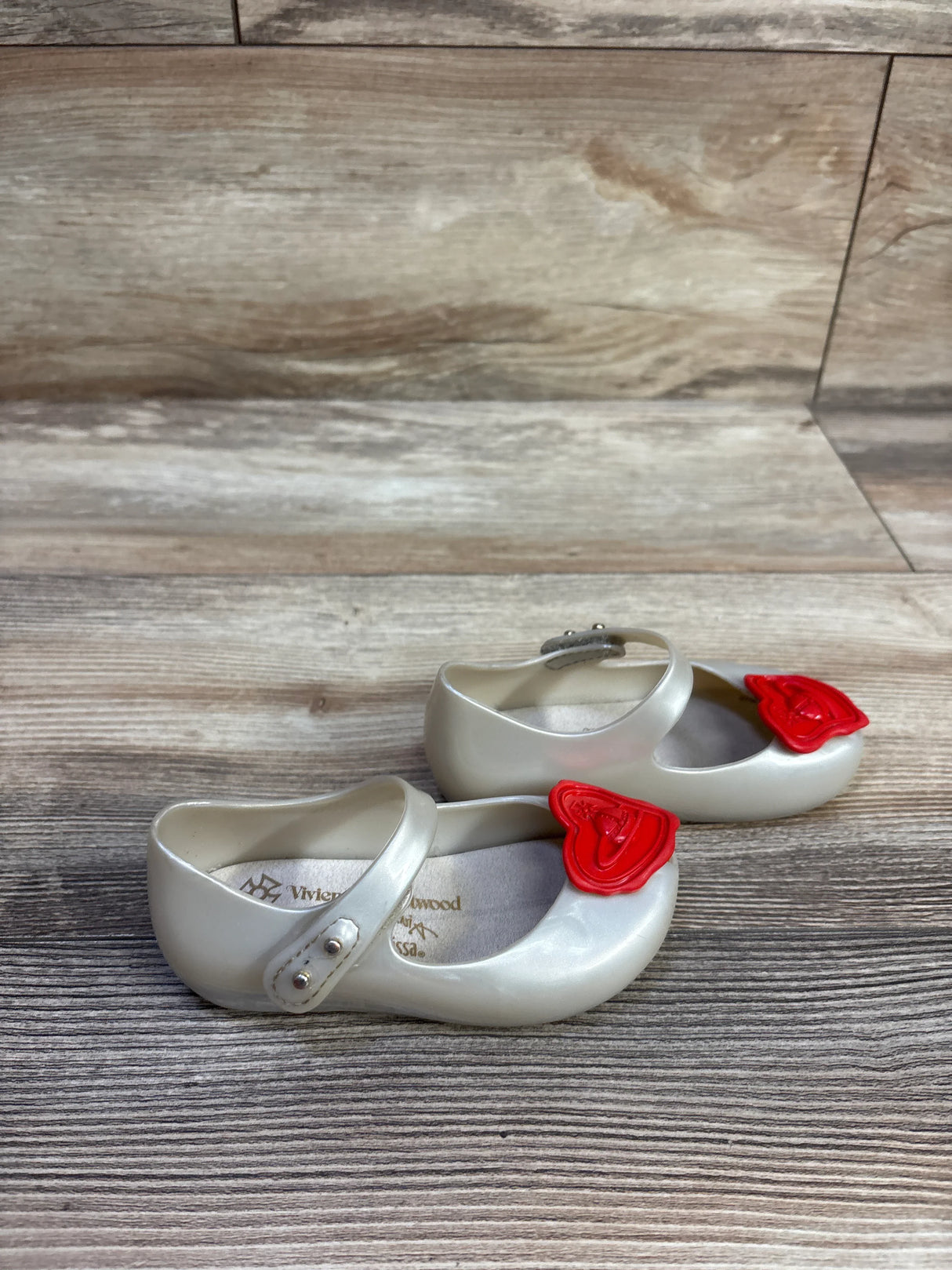 Side view of Vivienne Westwood x Mini Melissa Ultra Mary Jane shoes, pearlescent white with red heart bows on wood floor