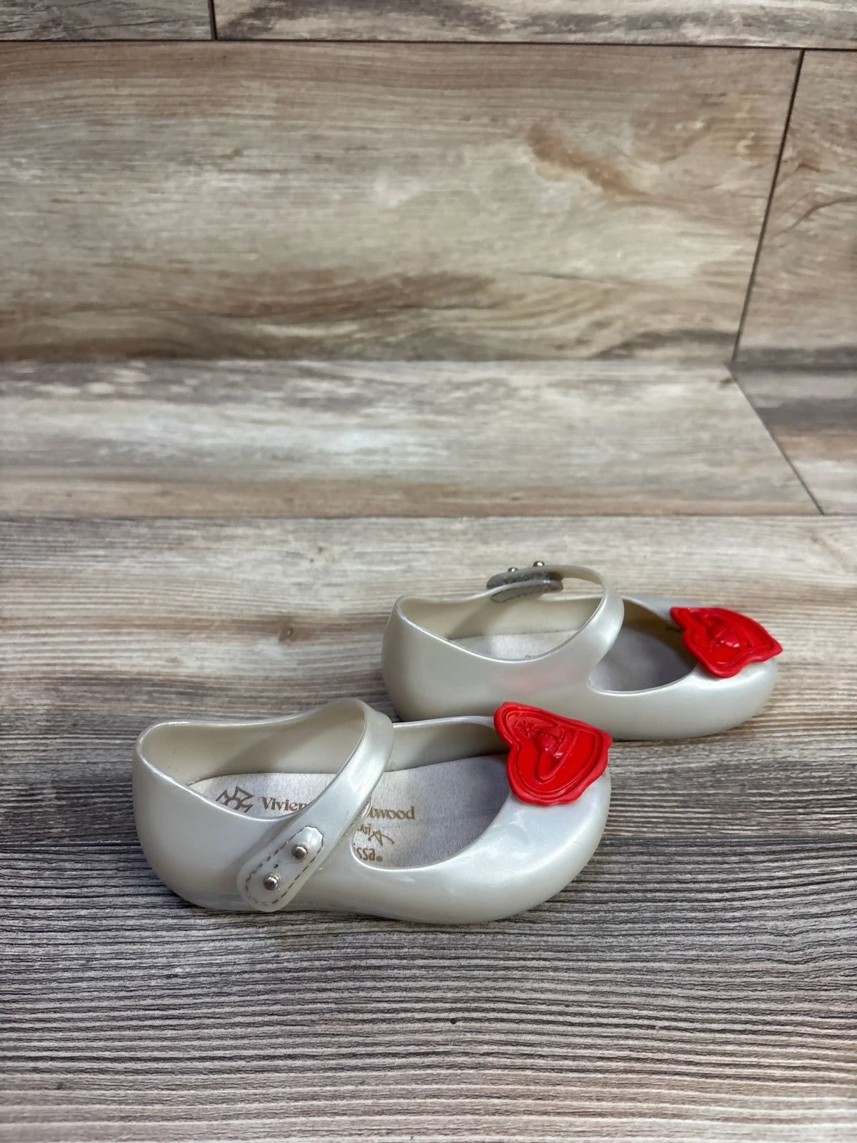 Side view of Vivienne Westwood x Mini Melissa Ultra Mary Jane shoes, pearlescent white with red heart bows on wood floor