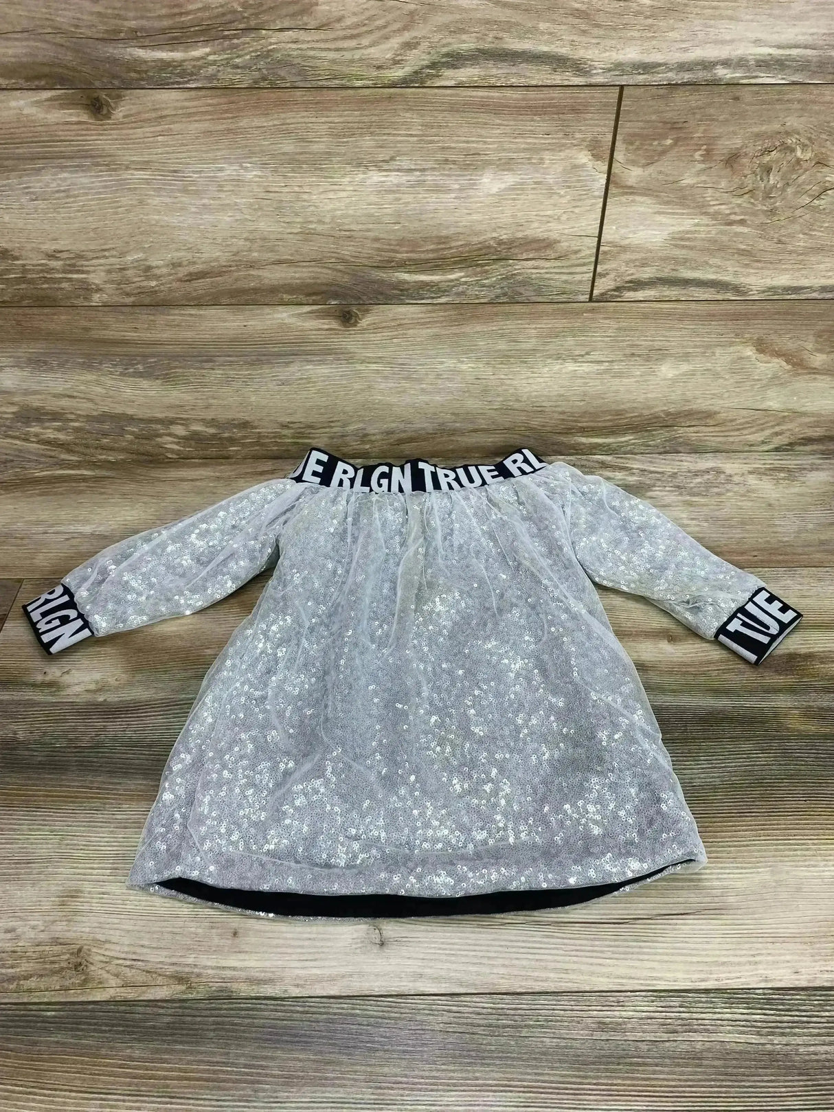 Flat lay of True Religion silver sequin dress with long sleeves and TRUE RLGN elastic neckline and cuffs on wood floor