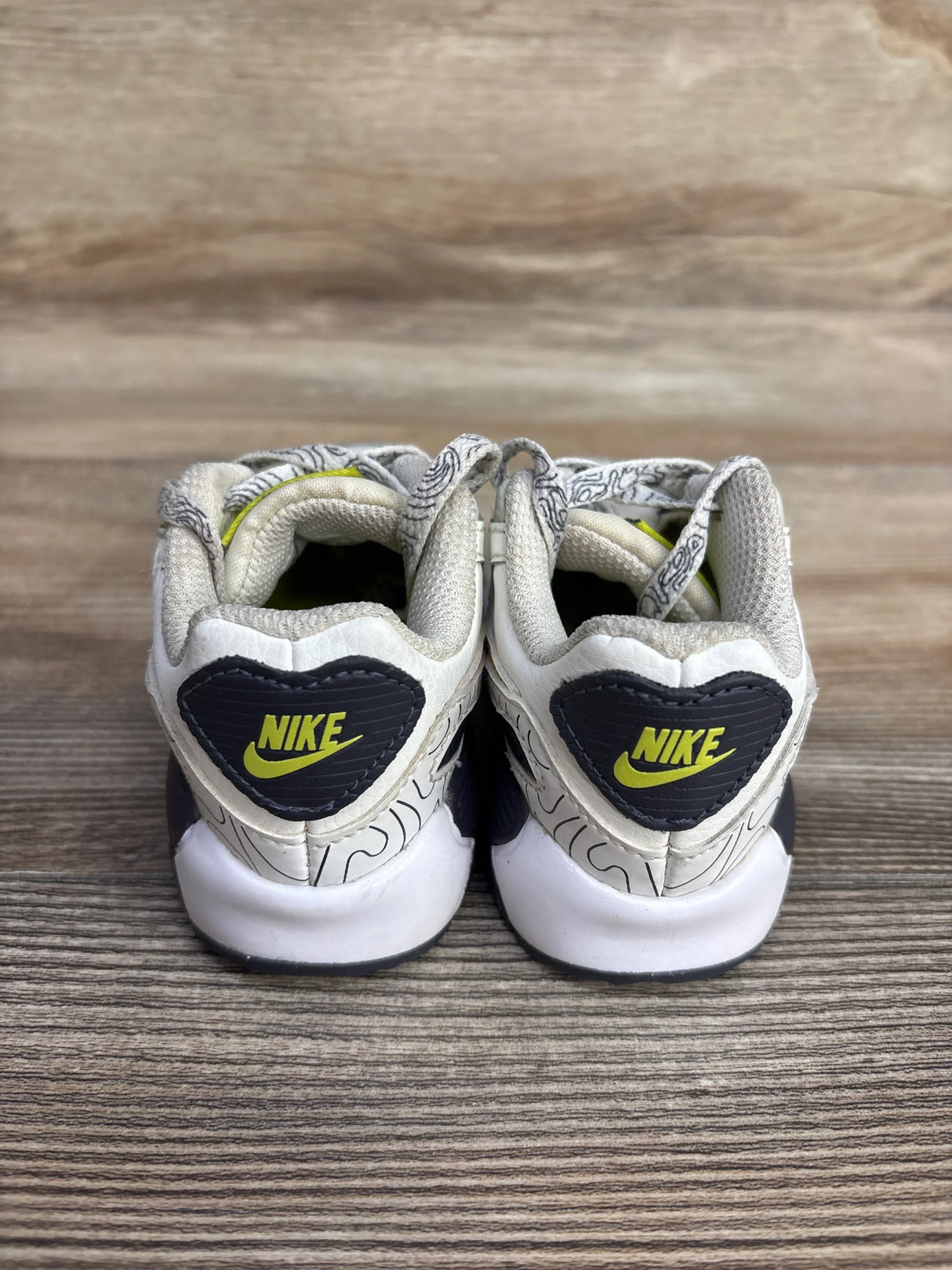 Rear view of Nike Air Max 90 beige sneakers size 4.5C showing black heart heel patches with yellow swoosh on wooden surface