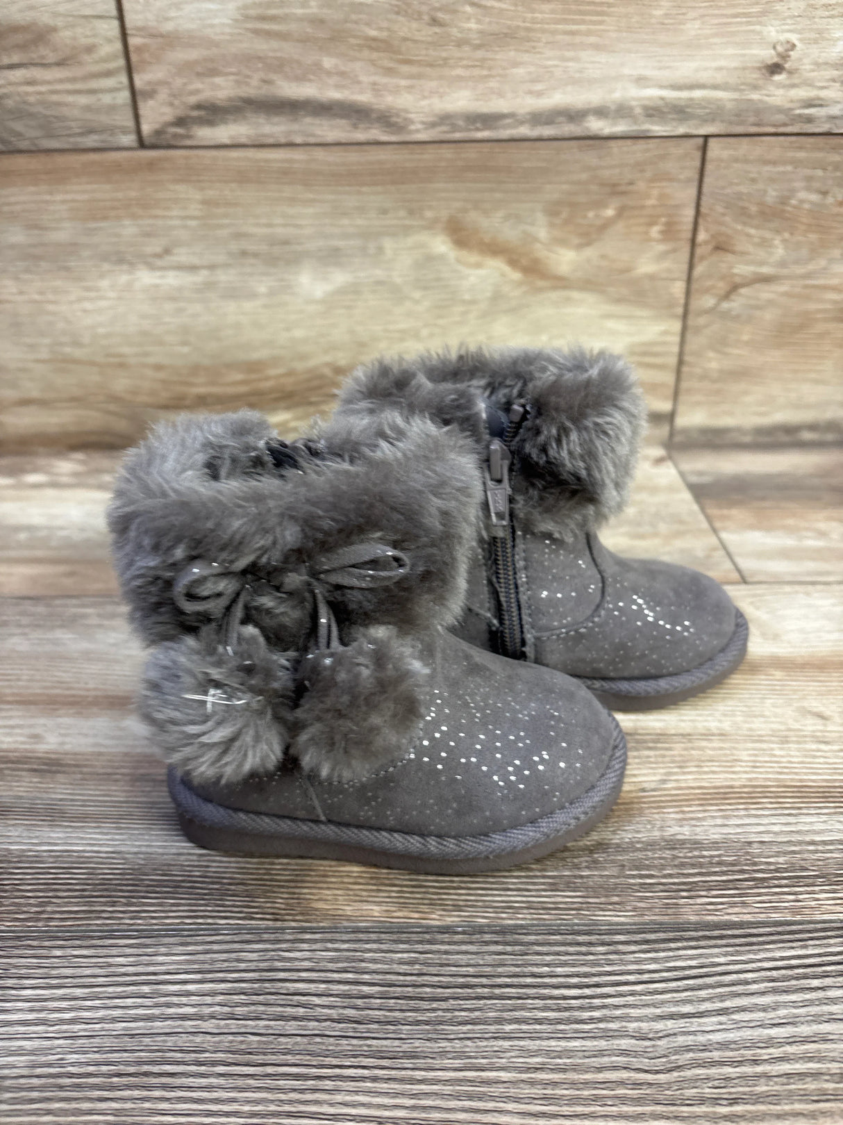 Three-quarter view of Jumping Beans Coco Easy Zip boots in grey with faux fur cuff, pom-pom ties and side zipper on wood