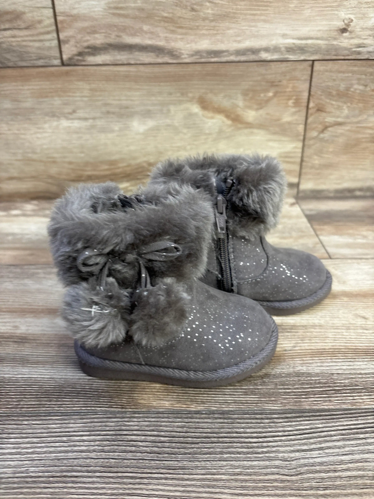 Three-quarter view of Jumping Beans Coco Easy Zip boots in grey with faux fur cuff, pom-pom ties and side zipper on wood