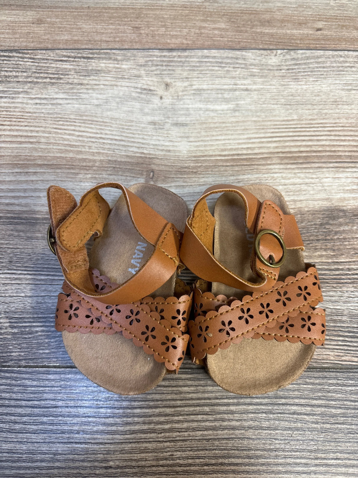 Top view of Old Navy perforated faux-leather baby sandals in beige with floral cutout straps and hook-and-loop closure on wood