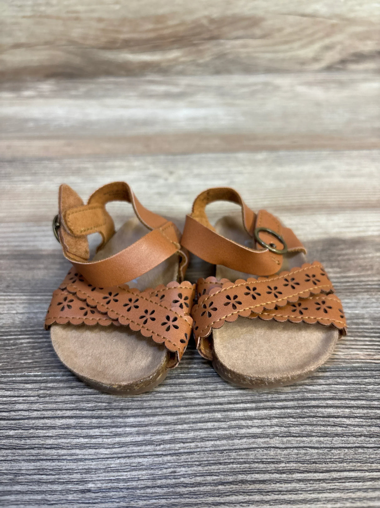 Front view of Old Navy baby sandals showing perforated scalloped floral straps on wood background