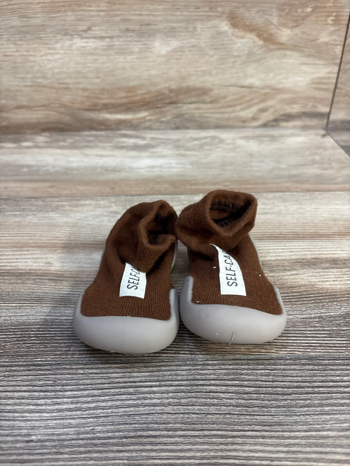 Front view of Tiny Explorings self-care sock shoes, brown size 2/3C, with light gray rounded soles on wood grain background