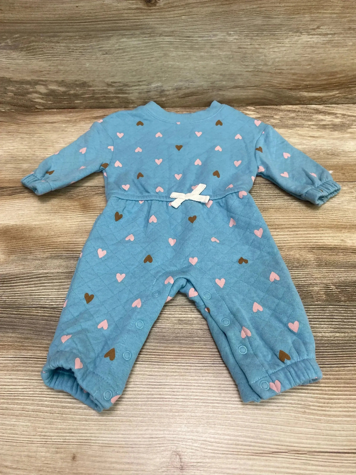 Front view of blue quilted baby coverall with heart print, white bow, snap leg closures and elastic cuffs on wood background