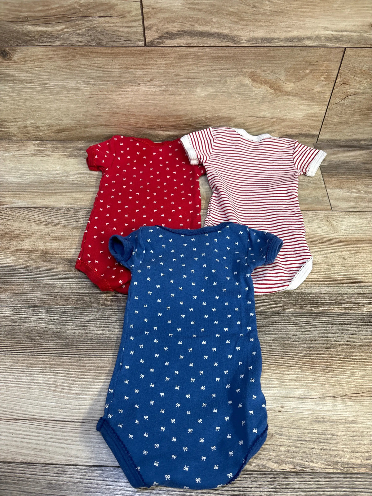Flat lay of three Petit Bateau short-sleeve bodysuits: navy with white bows, solid red, red-and-white stripe; 6m on wood floor