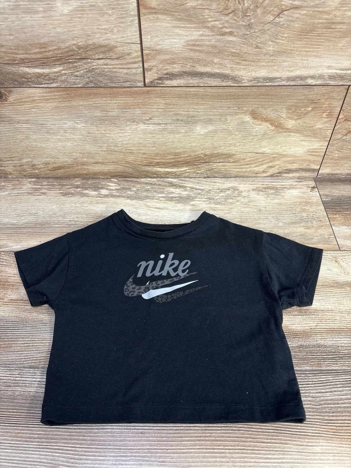 Front view of black toddler Nike tee showing grey logo and cheetah-print swoosh on wood background