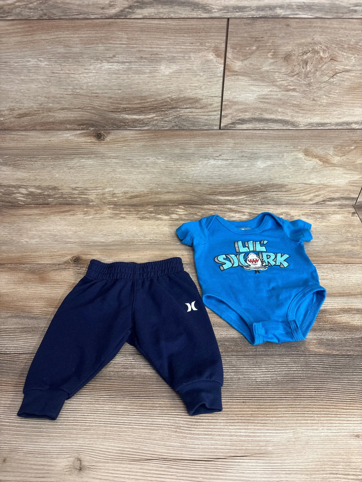 Flat lay of Hurley 2-piece baby set showing Lil Shark graphic on blue bodysuit and navy pants on wood floor, size 3m