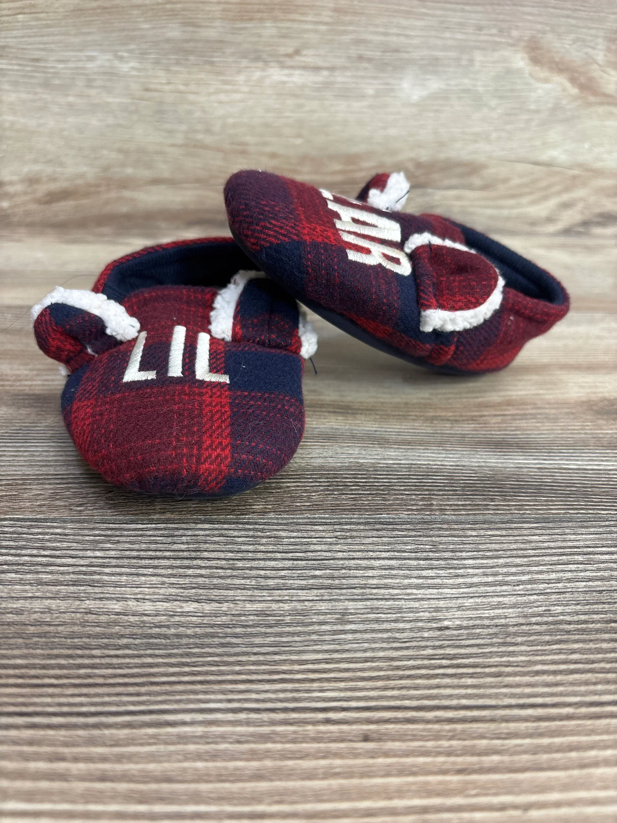 Red and navy plaid baby crib shoes with white text and faux fur trim on wooden background