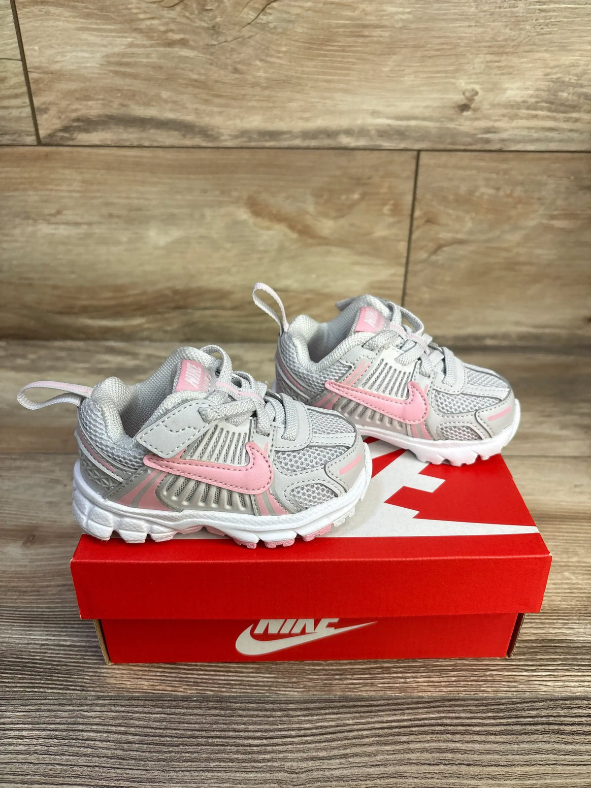 Front side view of toddler Nike Vomero 5 sneakers in grey with pink Swoosh and white sole on red Nike box over wood floor