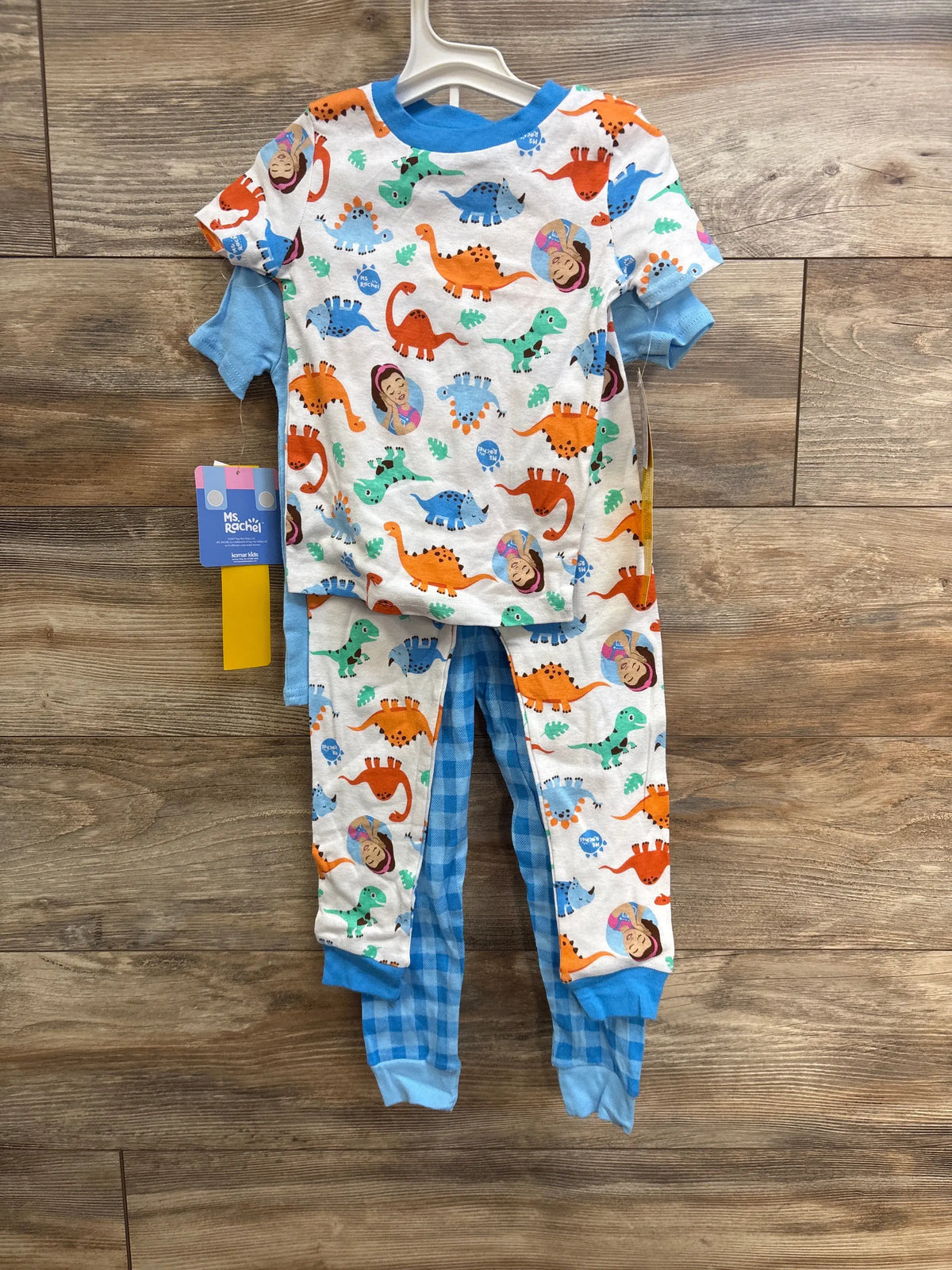 Flat lay of NEW Ms Rachel 4-piece snug-fit pajama set showcasing dinosaur-print tees and blue plaid pants on wood floor