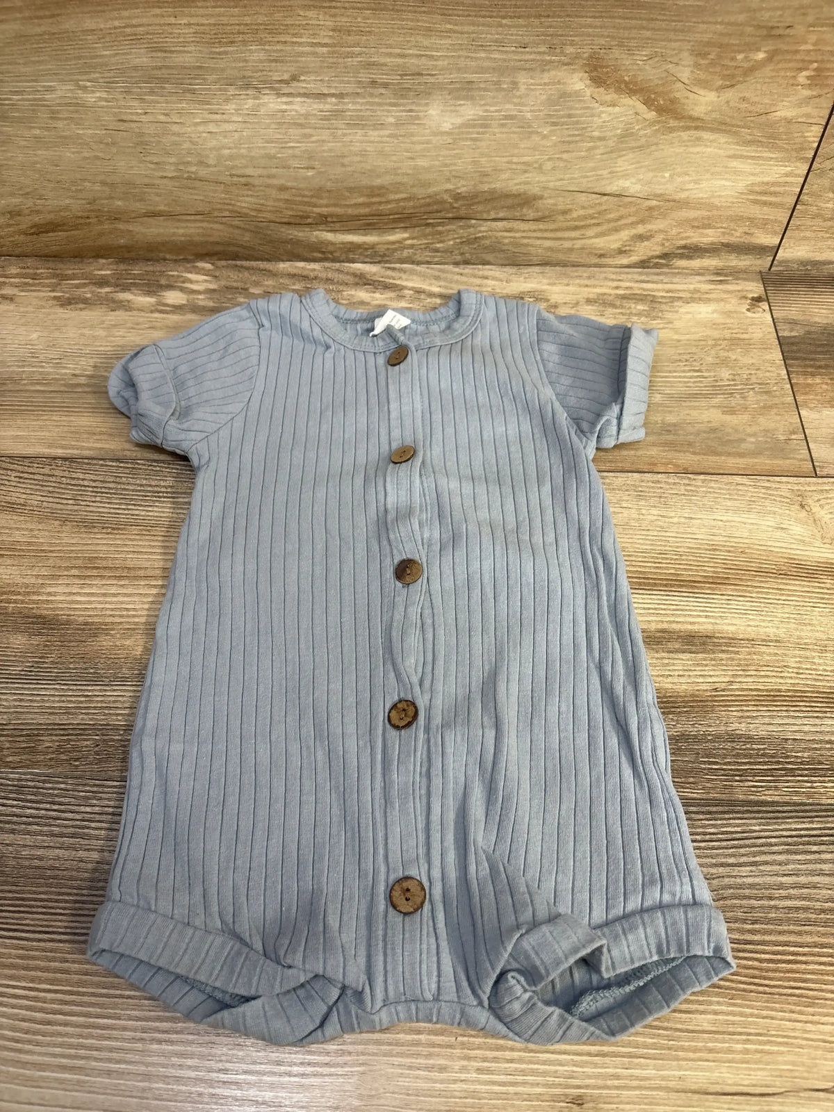 Front view of blue ribbed shortie romper showcasing button front and rolled leg openings on a wood floor
