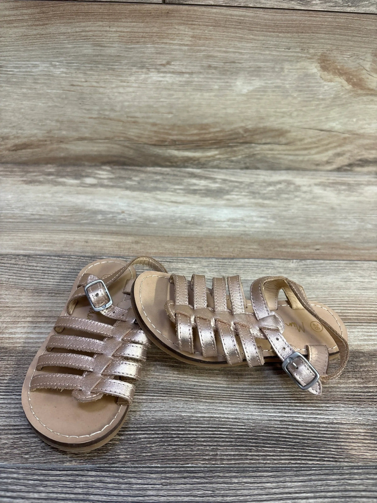 Angled view of Mini Boden toddler gladiator sandals in metallic gold showcasing braided straps and adjustable ankle buckle on wood plank background